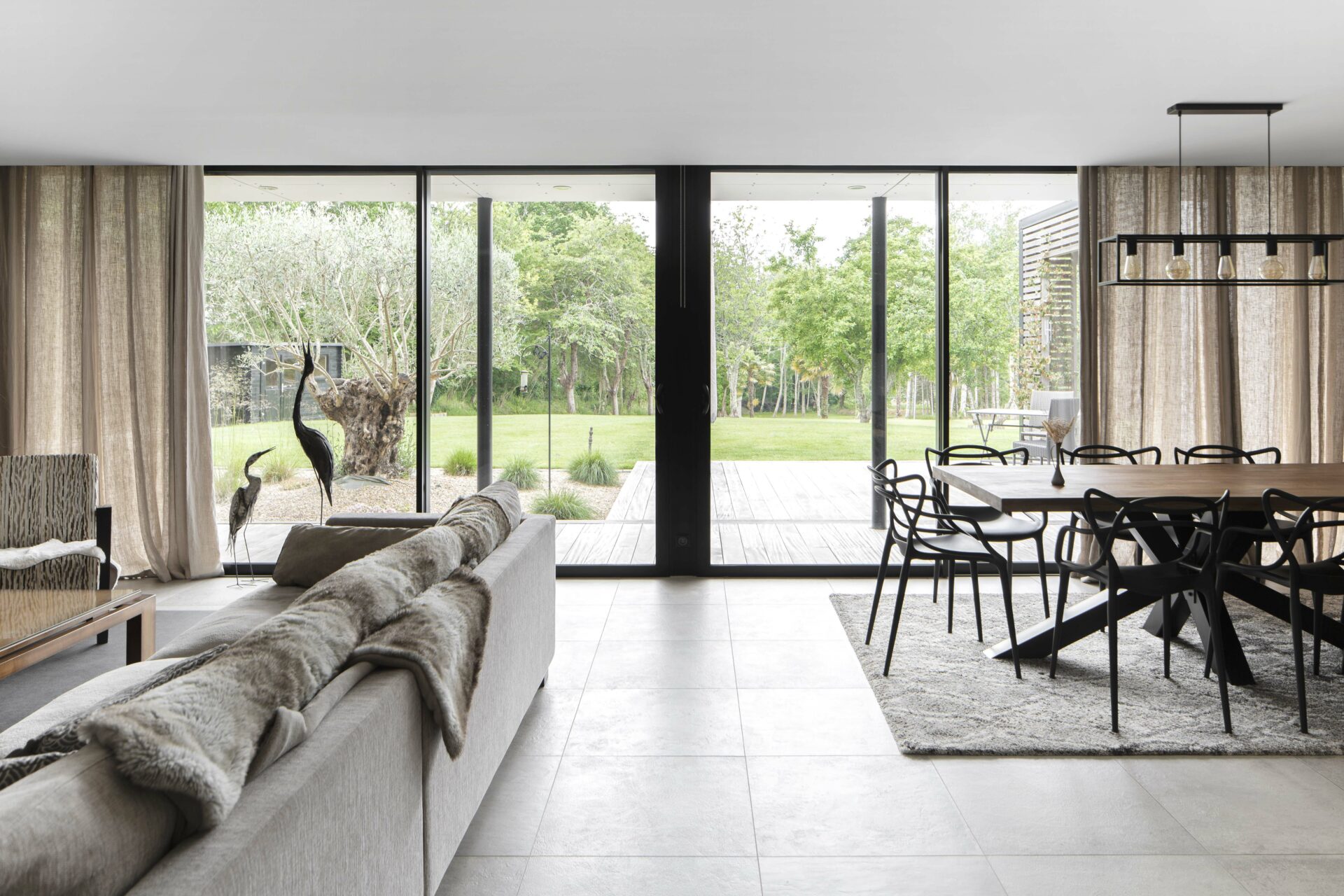 Grandes baies vitrées. Seuil encastré. Intérieur grande dalle de carrelage effet béton ciré. Terrasse en bois. Coursive sur le terrasse avec casquette en alumium blanc. Rideau en lin beige avec encastrement dans plafond.
